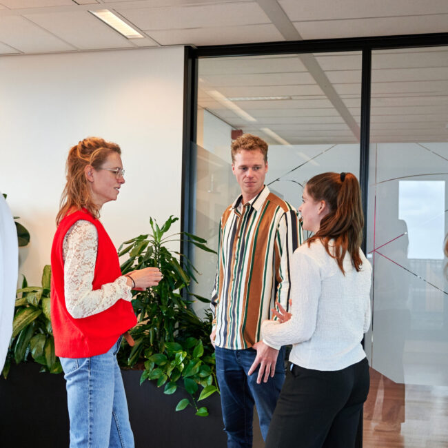 Collega's op kantoor – grote groep mensen zijn druk in gesprek met elkaar en er staan 2 mensen buiten de groep (zelfde context als vorige foto’s)>